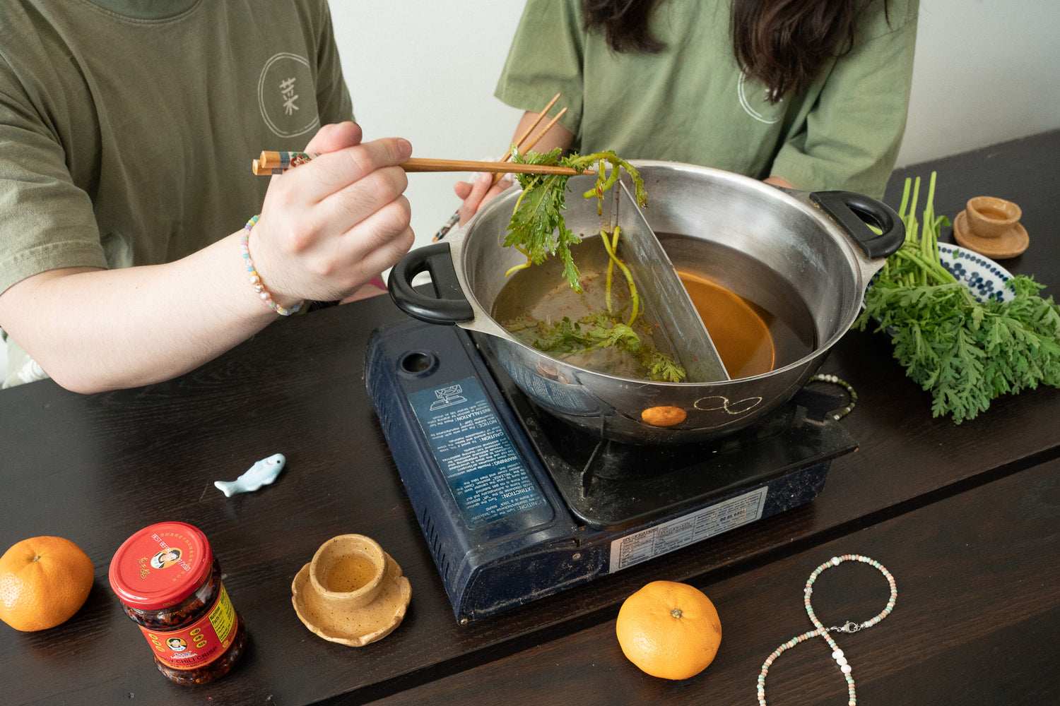 Two people wearing the same yellow green shirt sit around a wooden table. Only their chests can be seen in the photo. One hand is swirling chrysanthemum greens with chopsticks in the portable hot pot machine in front of them.