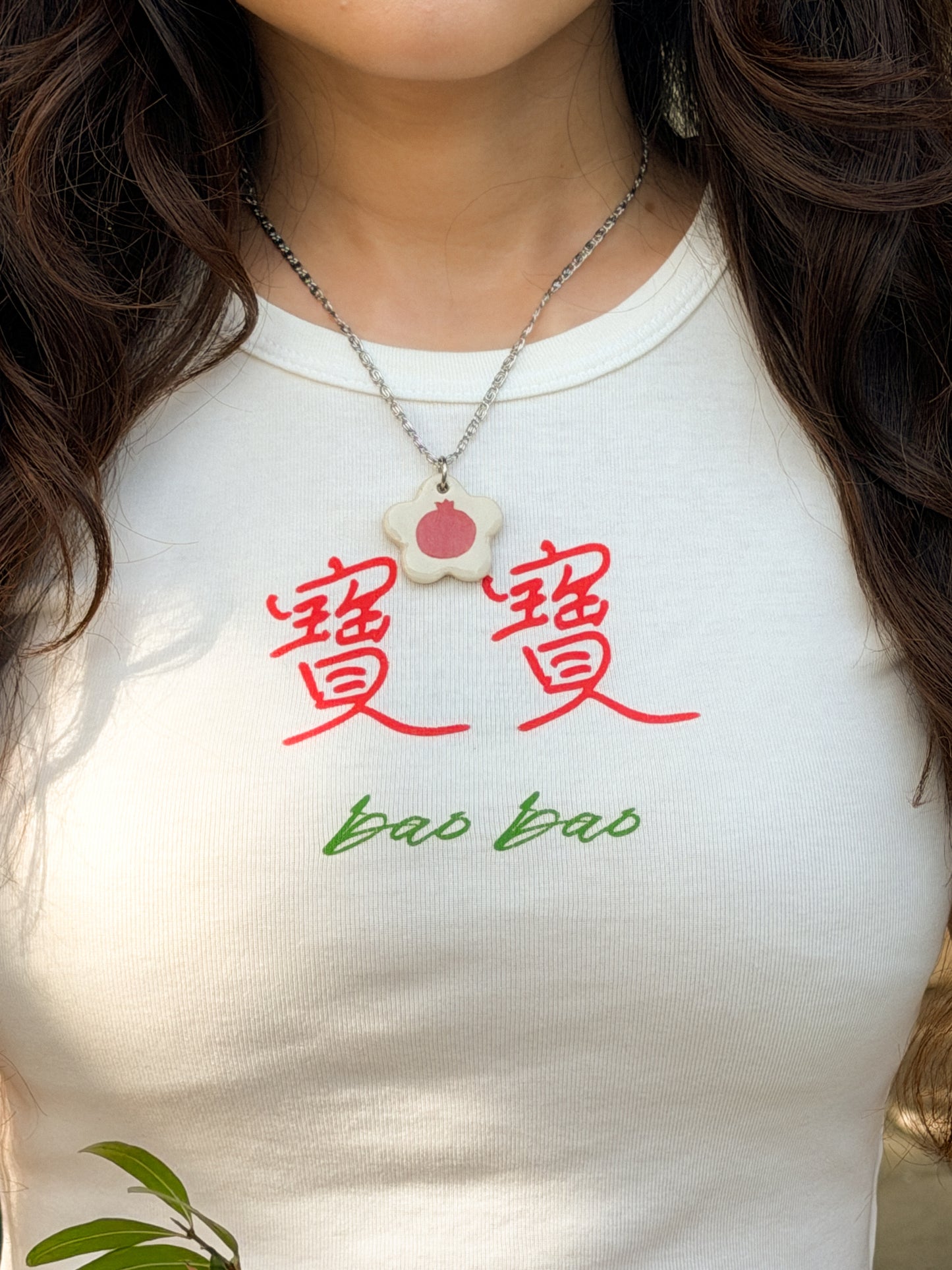 Close-up of person wearing a white baby tee with the Chinese words 寶寶 and "bao bao" on it and a necklace with a flower-shaped ceramic pendant featuring a pomegranate