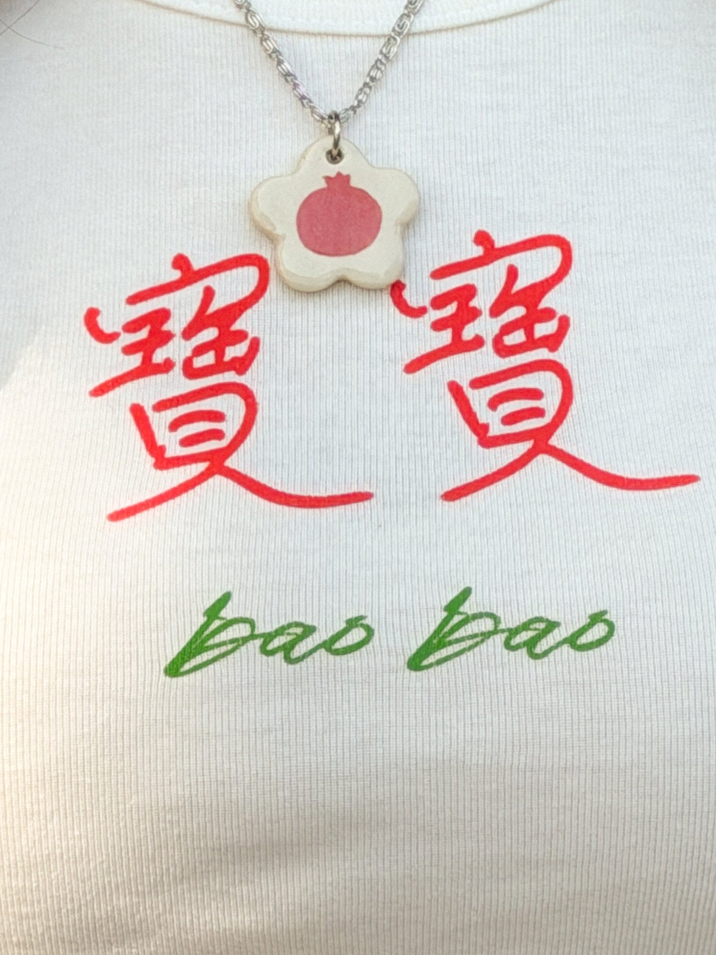 Close-up of person wearing a white baby tee with the Chinese words 寶寶 and "bao bao" on it and a necklace with a flower-shaped ceramic pendant featuring a pomegranate