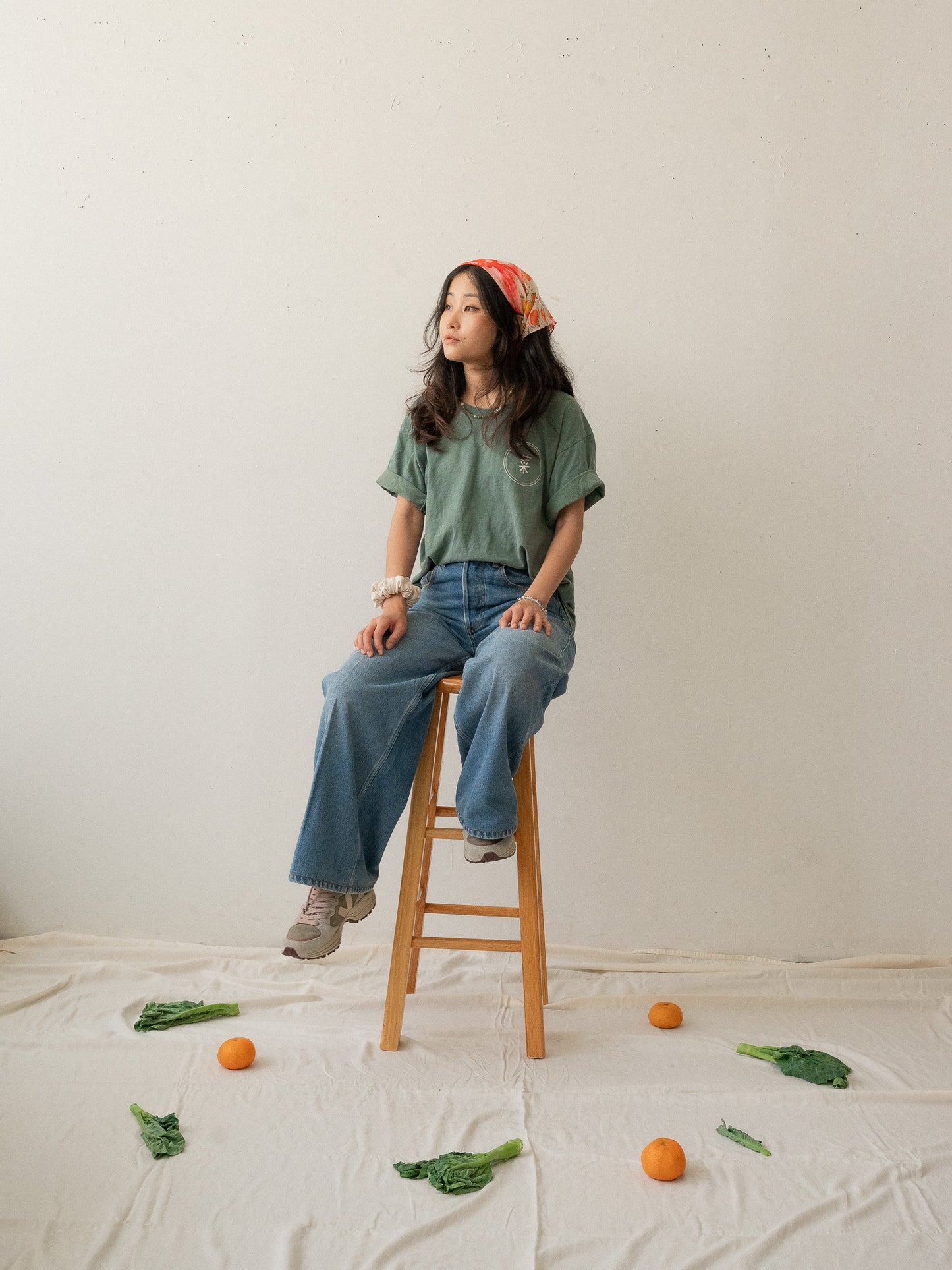 A Chinese Vietnamese woman with long hair wearing a green t-shirt and blue jeans sits on a wooden chair with her face turned slightly to the side and looking into the distance. On the floor are tangerines and Chinese broccoli on top of a white sheet.