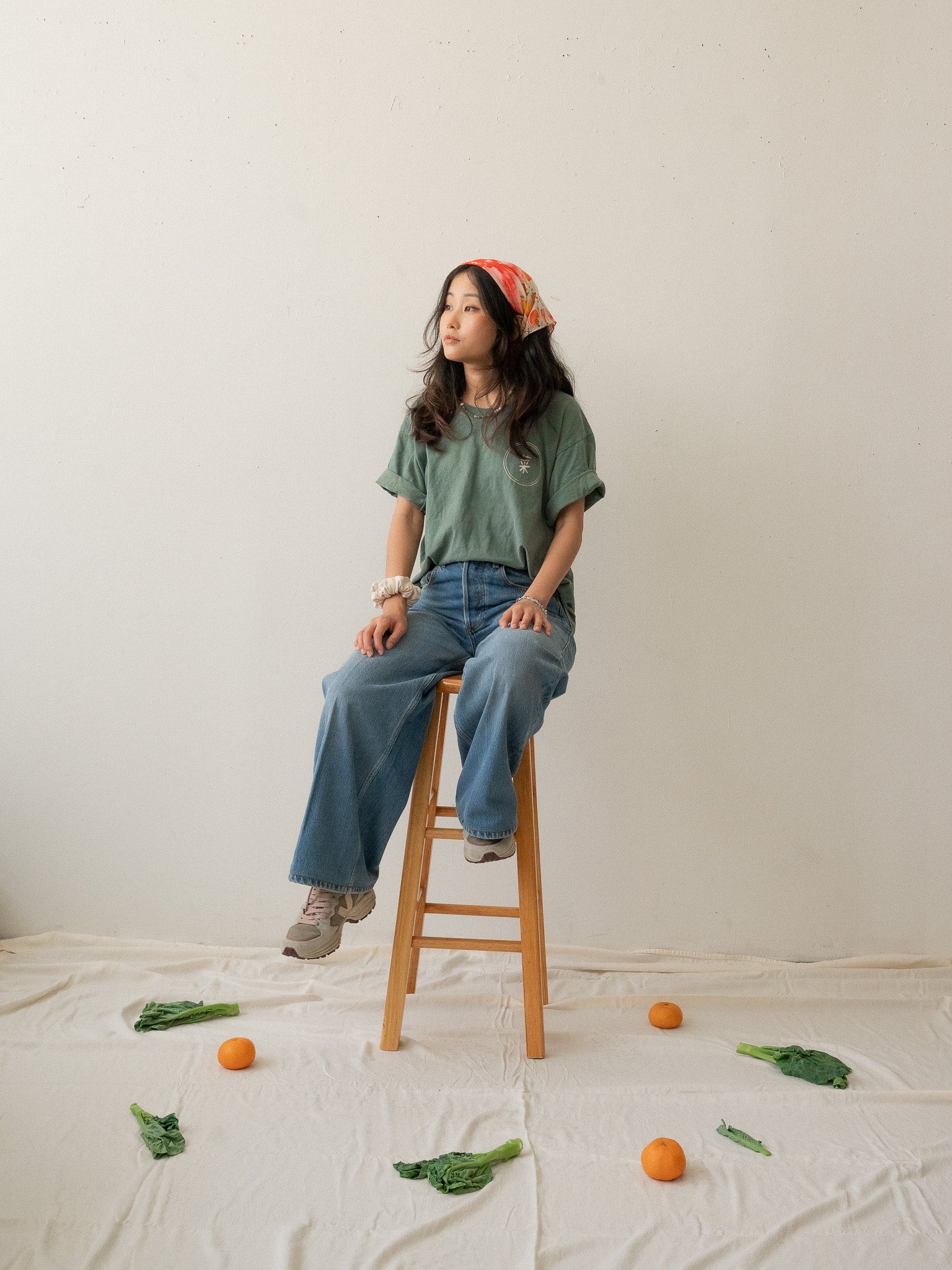 A Chinese Vietnamese woman with long hair wearing a green t-shirt and blue jeans sits on a wooden chair with her face turned slightly to the side and looking into the distance. On the floor are tangerines and Chinese broccoli on top of a white sheet.