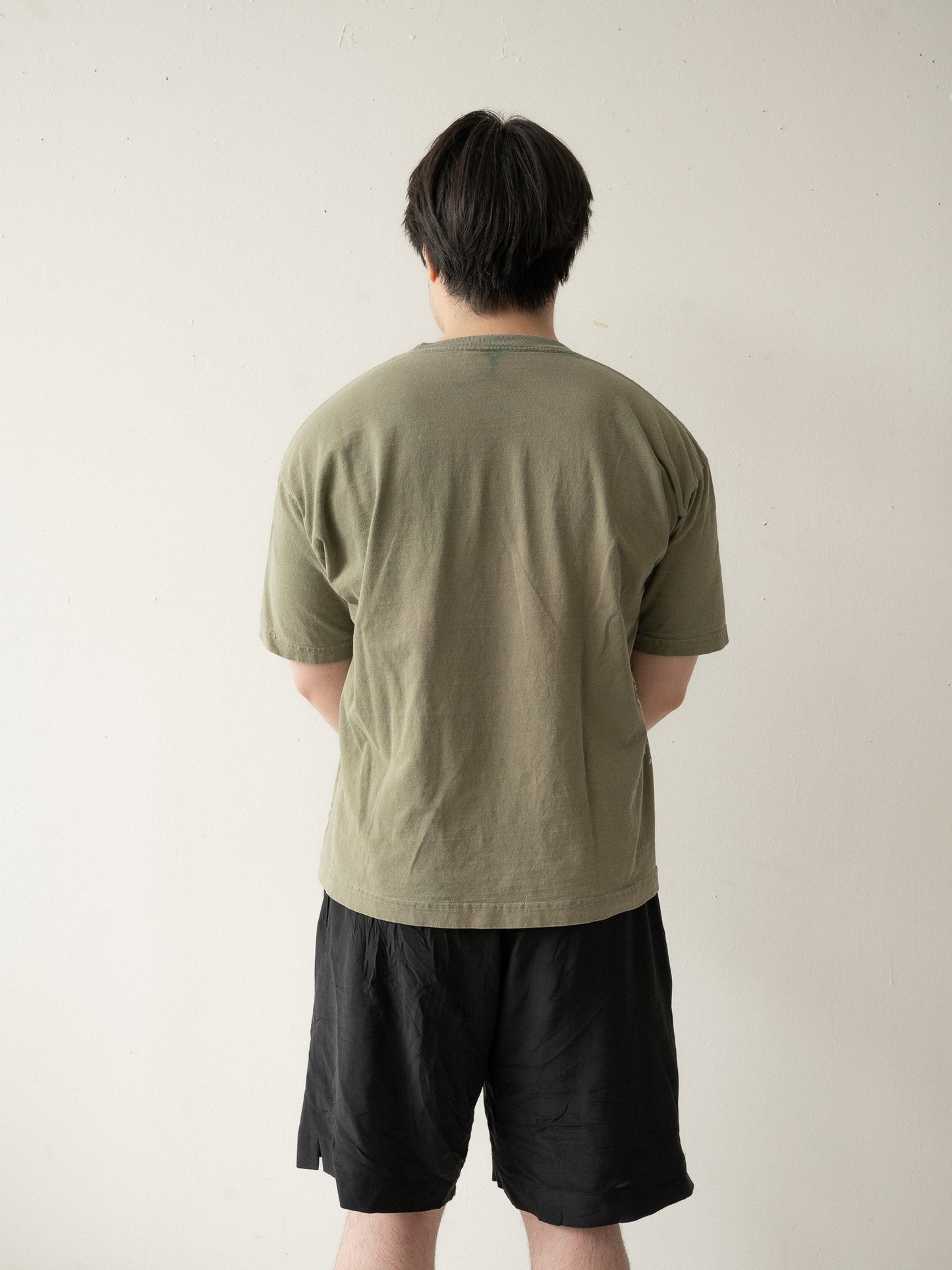 A Chinese man wearing a yellow brownish green t-shirt and black shorts stands with his back to the camera.
