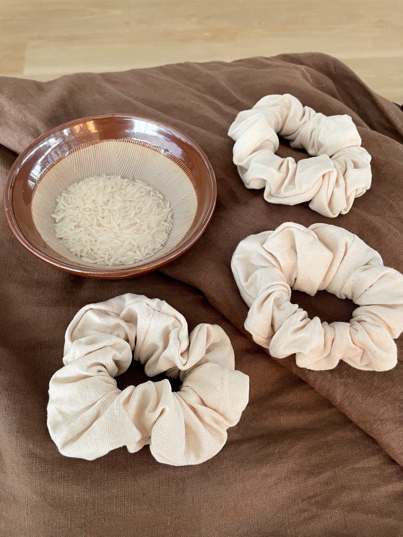 Three off white scrunchies lay flat on a brown cloth next to a ceramic bowl of uncooked rice.