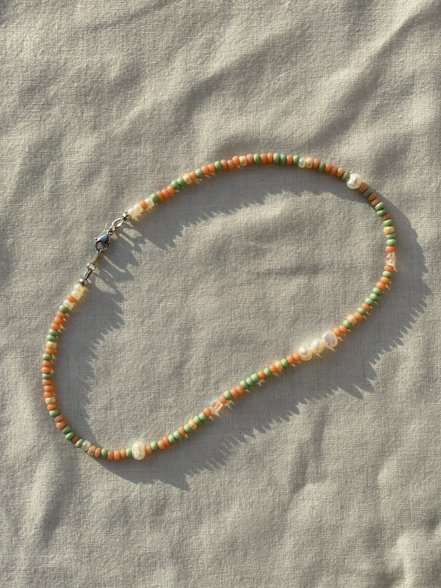 A necklace made of Czech glass beads, white freshwater pearls, and stainless steel lay flat on a white piece of cloth in the sunlight.