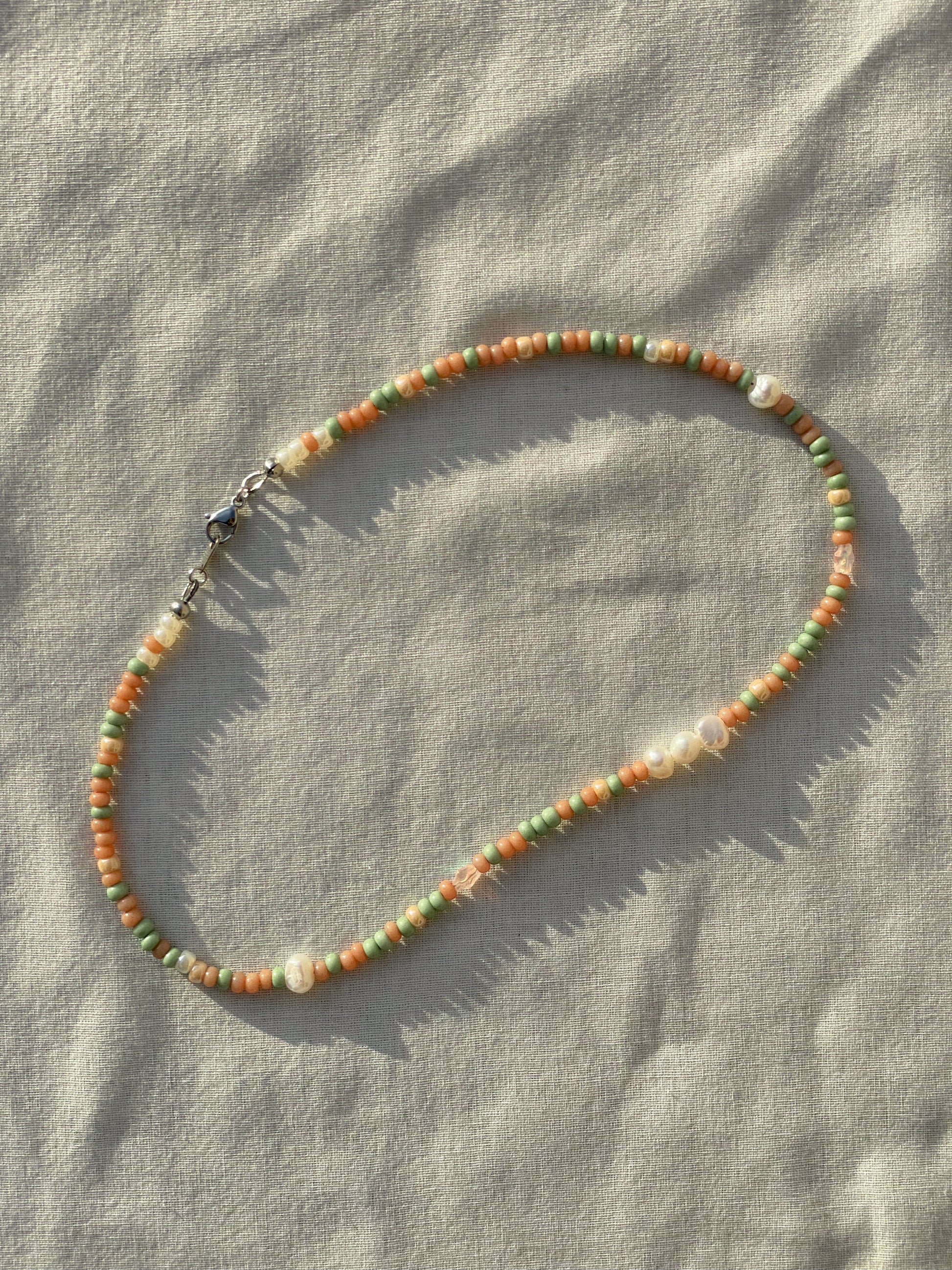 A necklace made of Czech glass beads, white freshwater pearls, and stainless steel lay flat on a white piece of cloth in the sunlight.