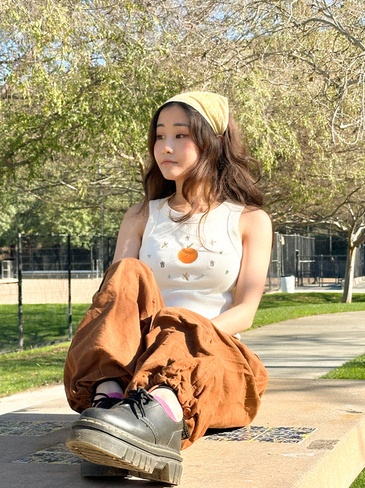 Chinese Vietnamese girl wearing a tank top, brown pants, and yellow bandana sits in a table looking to the side
