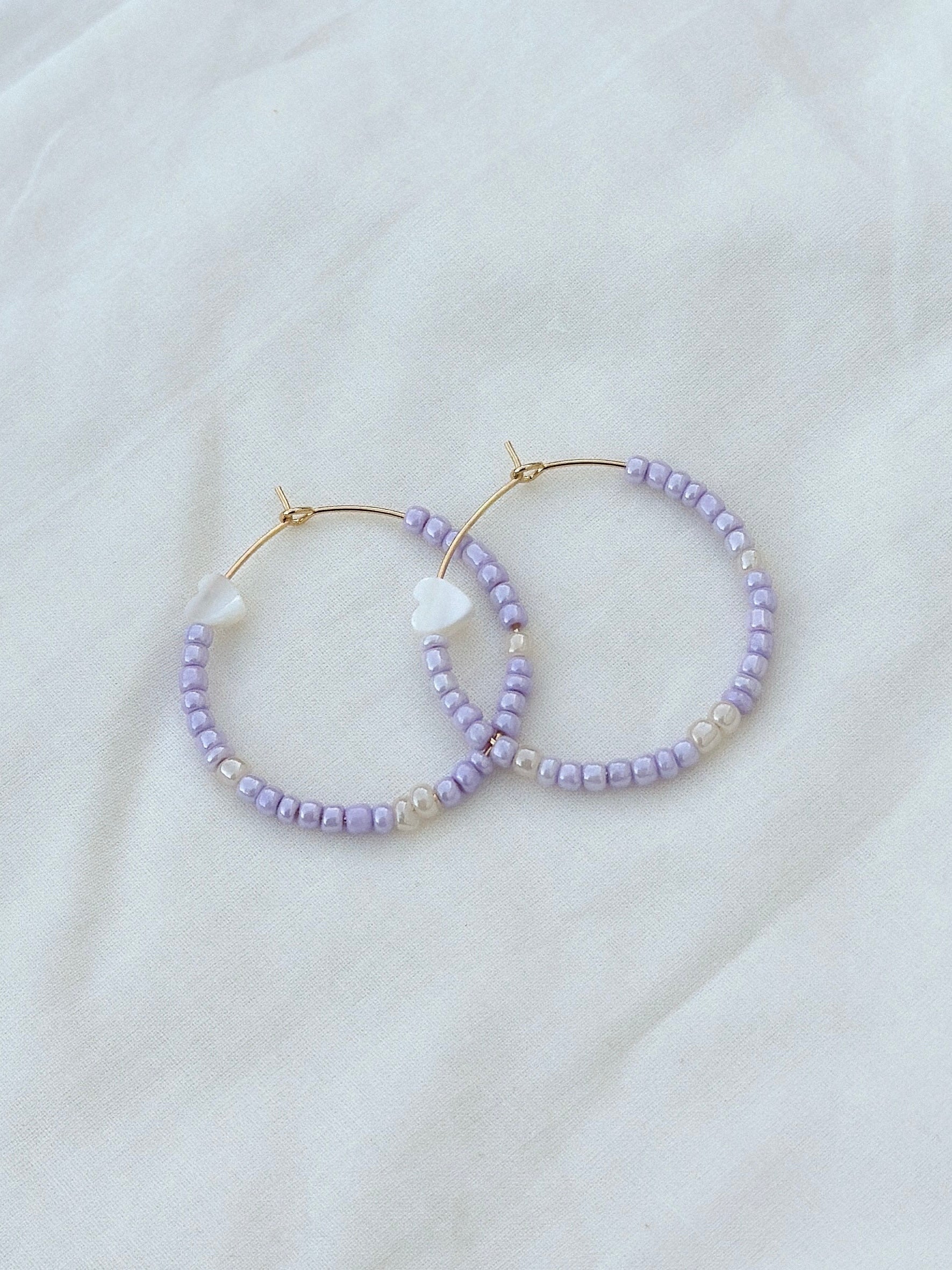 a pair of gold stainless steel earrings strung with shiny purple and white beads and heart-shaped mother of pearls lay flat on top of a piece of fabric