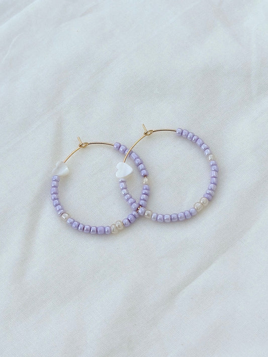 a pair of gold stainless steel earrings strung with shiny purple and white beads and heart-shaped mother of pearls lay flat on top of a piece of fabric