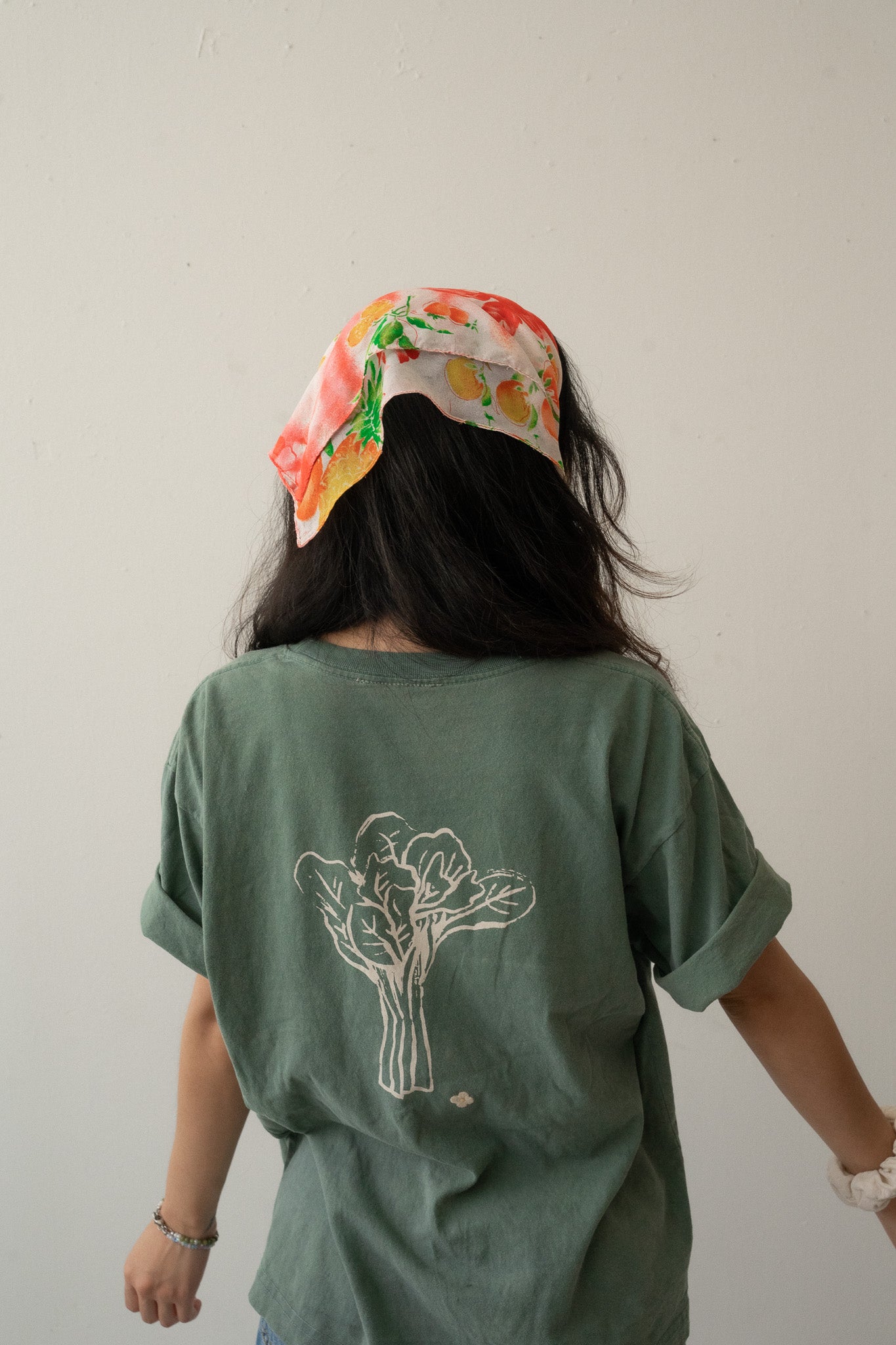 A Chinese Vietnamese woman wearing a green t-shirt with an illustration of a big Chinese broccoli and small embroidered white flower and blue jeans stands with her back to the camera. 