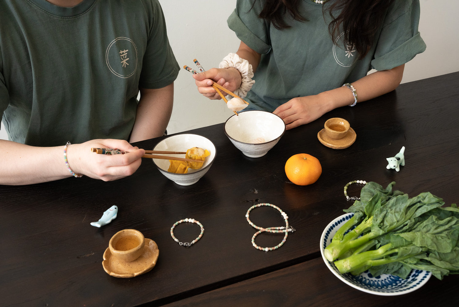 Two people wearing the same dark green shirt sit around a wooden table. Only their chests can be seen in the photo. They’re holding chopsticks. There are bowls of dim sum, tea cups, and a plate of Chinese broccoli on the table.