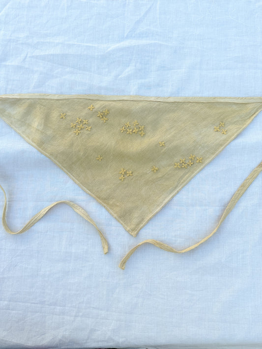 A muted yellow triangle-shaped bandana with two long straps and habe embroidered flowers lay on a white piece of cloth