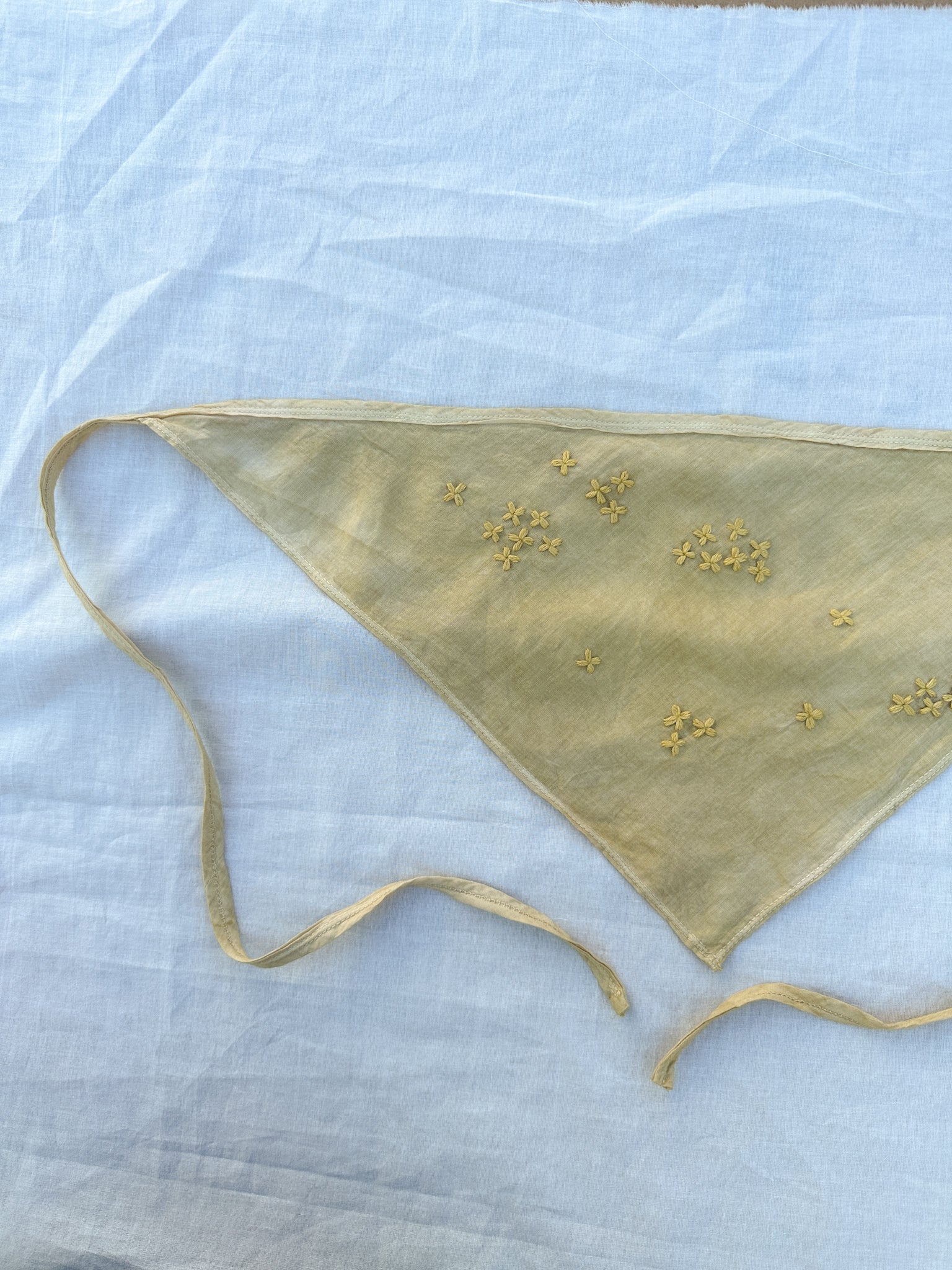 A muted yellow triangle-shaped bandana with two long straps and habe embroidered flowers lay on a white piece of cloth