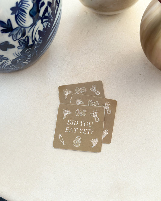 a few light brown stickers with the words “did you eat yet?” and illustrations of veggies lay flat on ceramic next to two vases