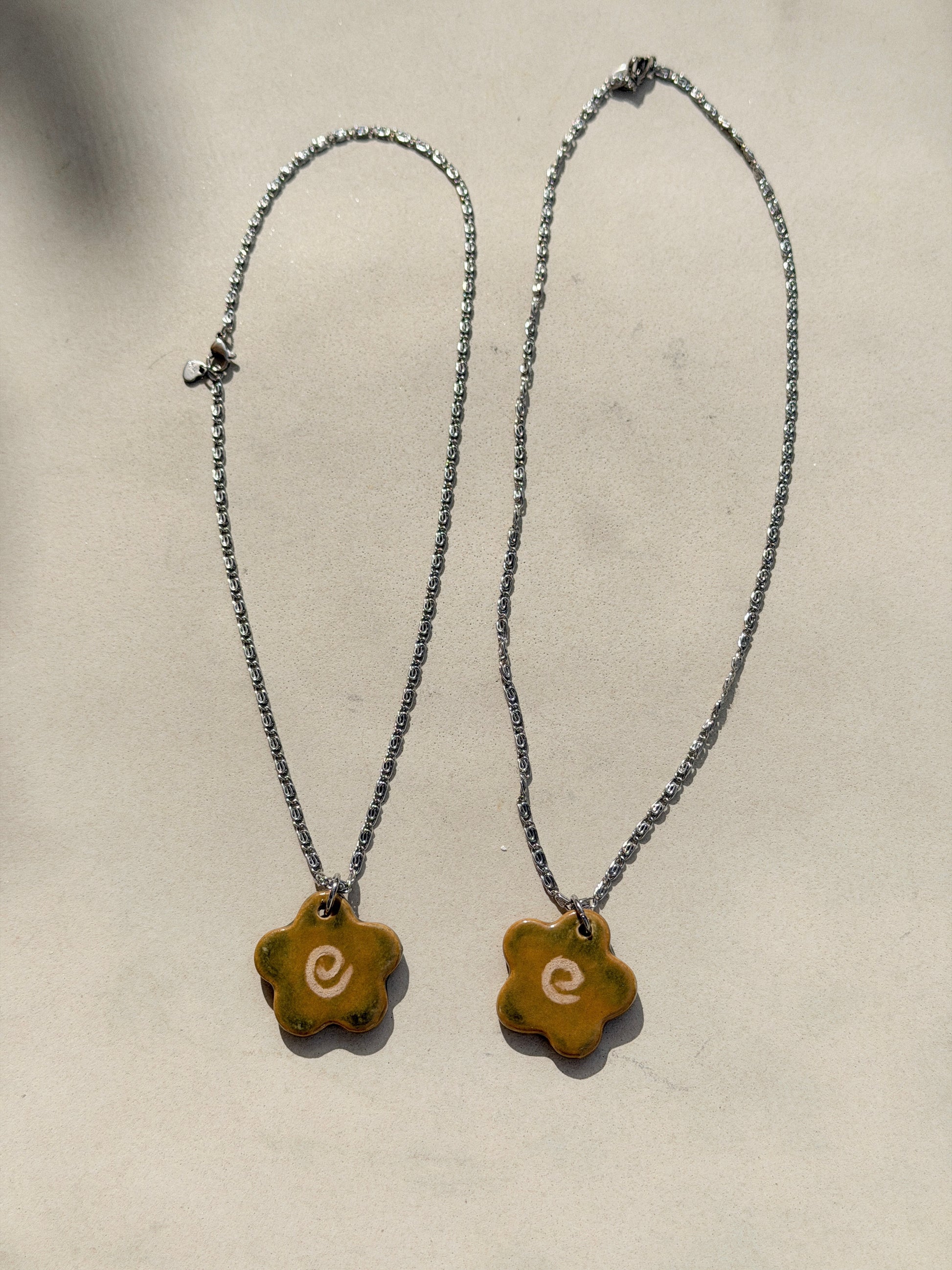 Full view of a stainless steel necklace with a glossy floral shaped ceramic pendant that is greenish yellow brown in color and has a hand-drawn cream swirl in the center