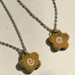 Partial view of two stainless steel necklaces with a glossy floral shaped ceramic pendant that is greenish yellow brown in color and has a hand-drawn cream swirl in the center