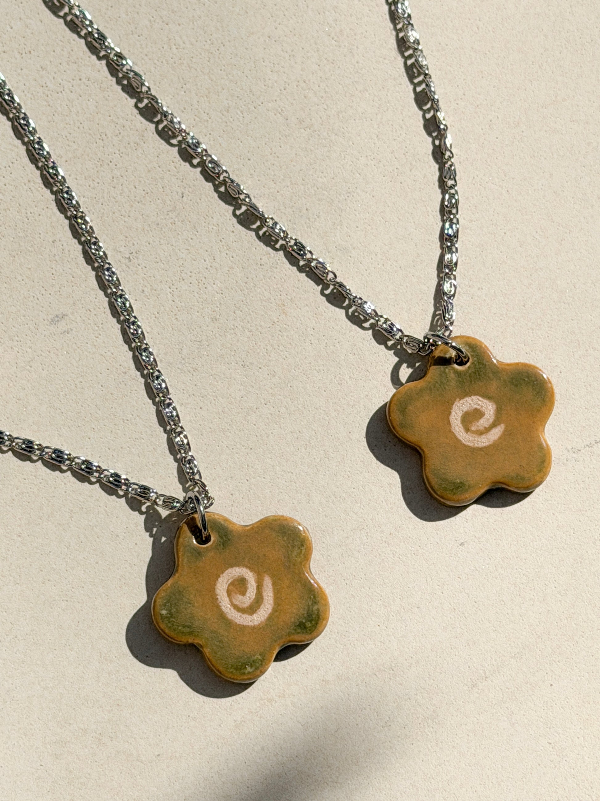 Partial view of two stainless steel necklaces with a glossy floral shaped ceramic pendant that is greenish yellow brown in color and has a hand-drawn cream swirl in the center