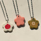 Partial view of three necklaces under the sun, each one featuring a stainless steel chain made of tiny swirls and a glossy floral-shaped ceramic charm with different designs