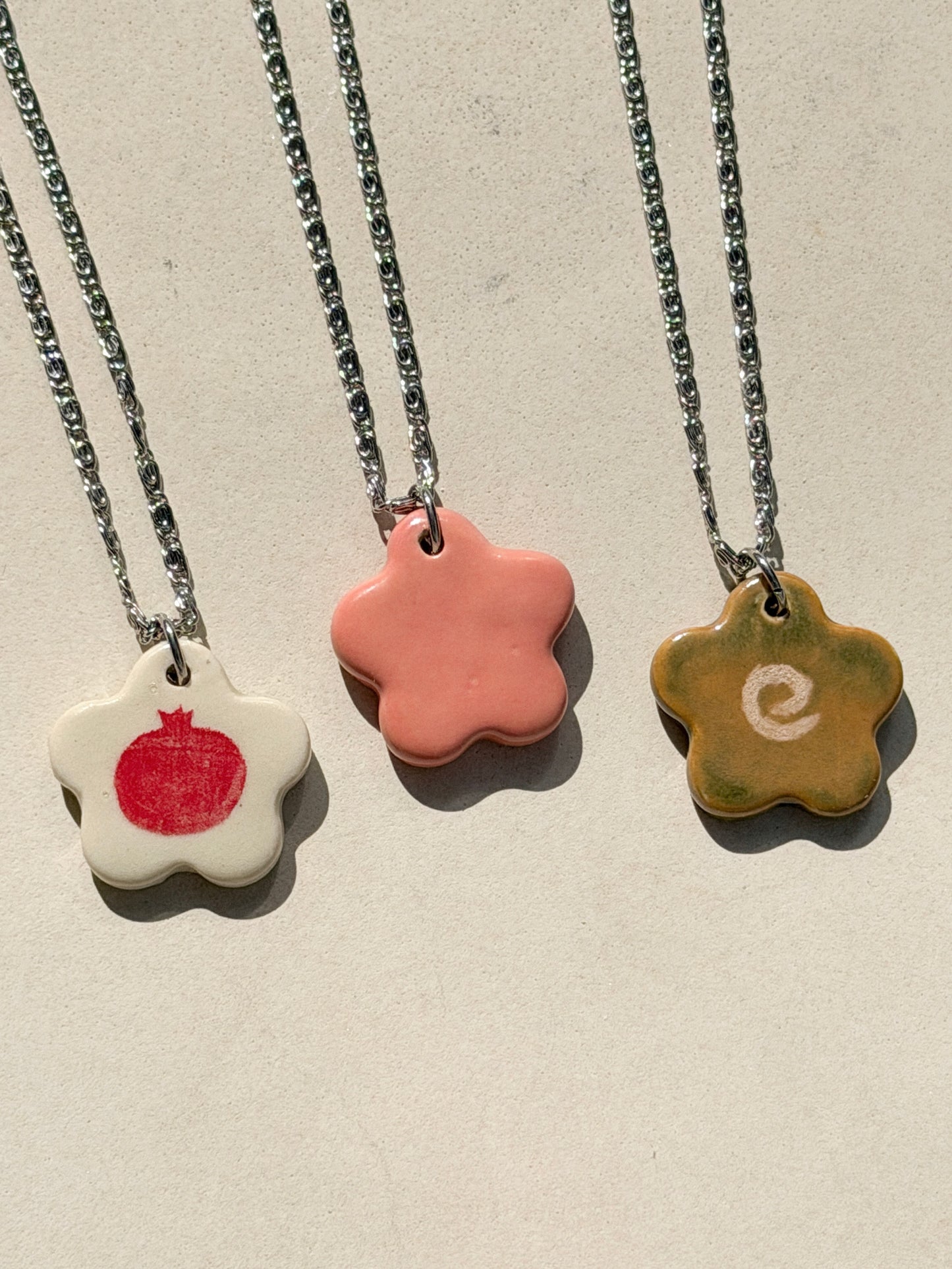 Partial view of three necklaces under the sun, each one featuring a stainless steel chain made of tiny swirls and a glossy floral-shaped ceramic charm with different designs