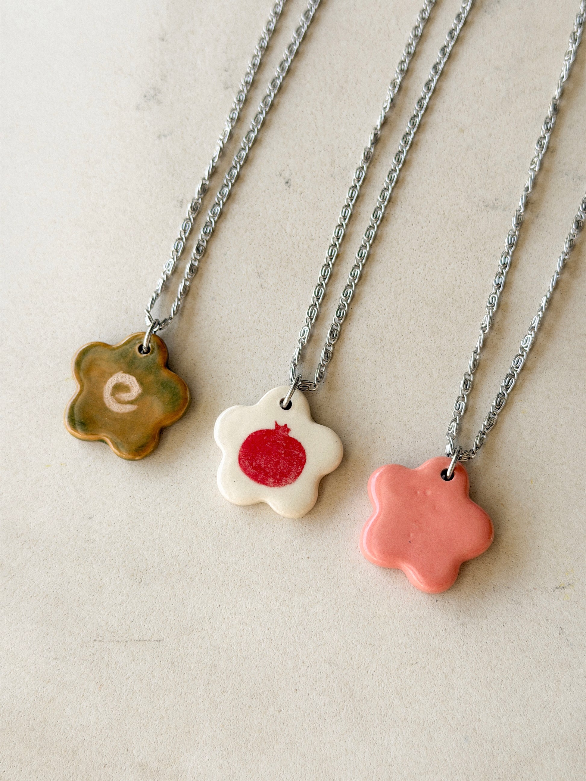 Partial view of three necklaces, each one featuring a stainless steel chain made of tiny swirls and a glossy floral-shaped ceramic charm with different designs
