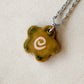 Partial view of a stainless steel necklace with a glossy floral shaped ceramic pendant that is greenish yellow brown in color and has a hand-drawn cream swirl in the center