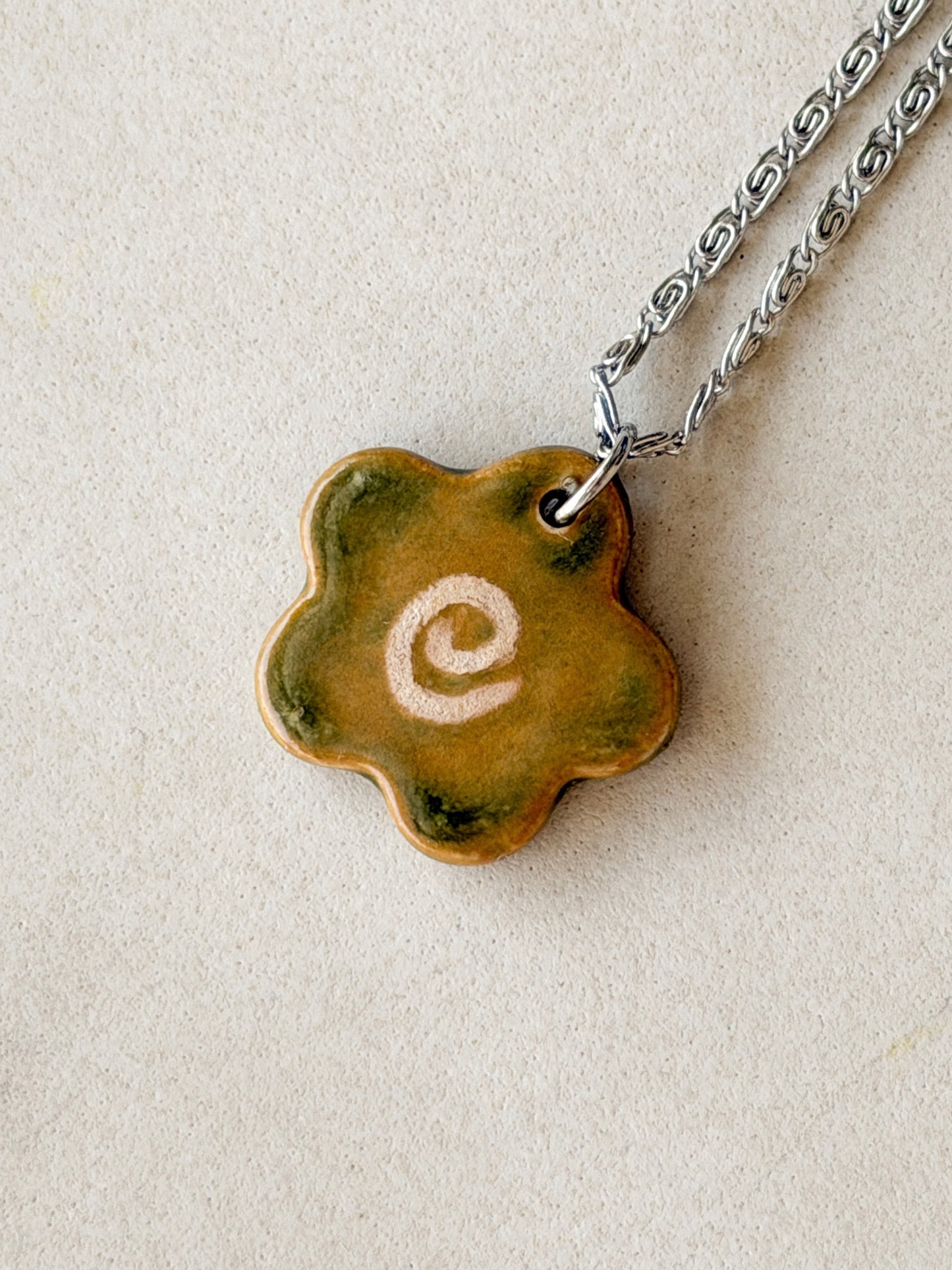 Partial view of a stainless steel necklace with a glossy floral shaped ceramic pendant that is greenish yellow brown in color and has a hand-drawn cream swirl in the center