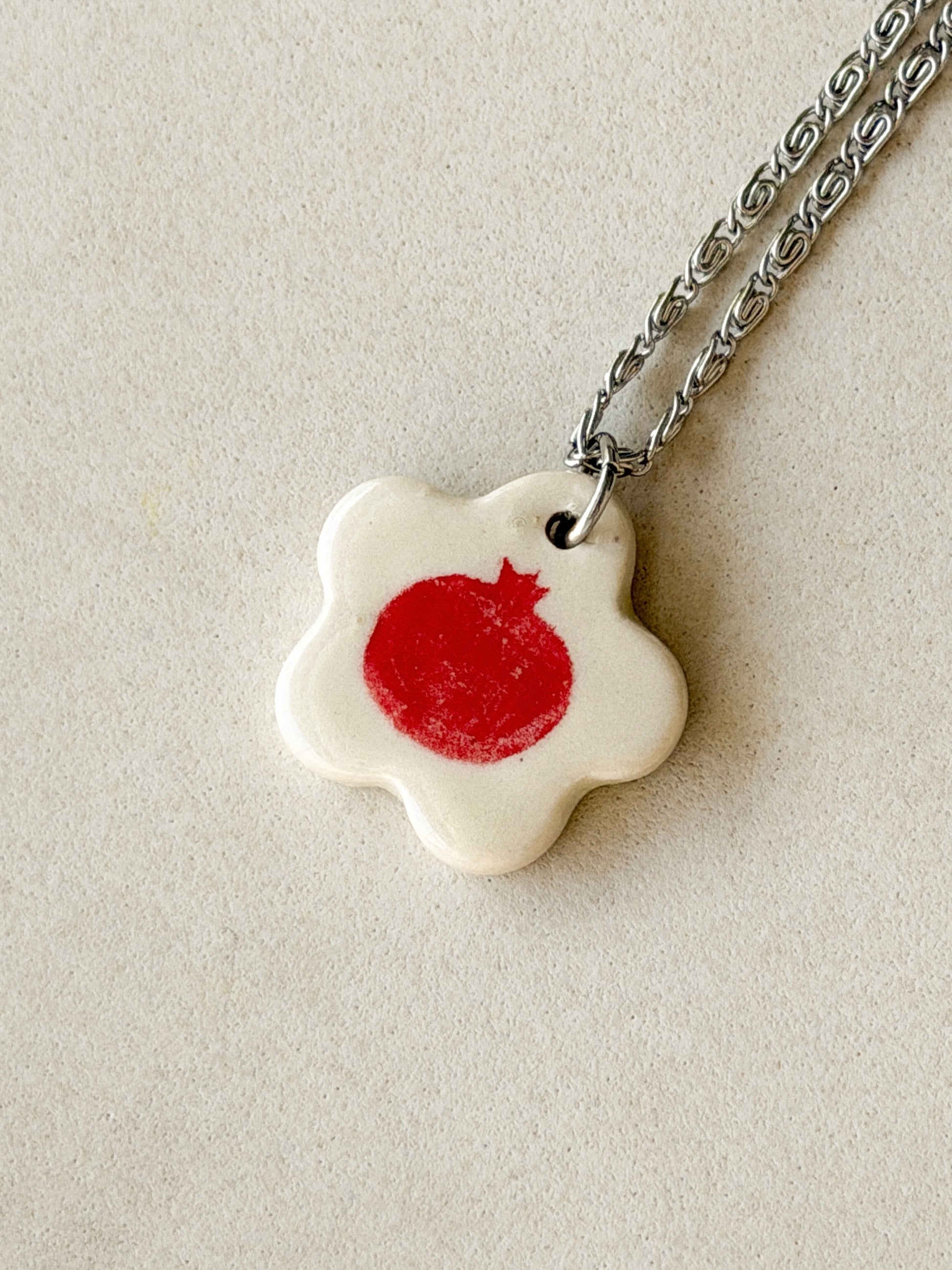 Stainless steel necklace with an off-white floral-shaped ceramic pendant featuring a distressed red pomegranate in the center