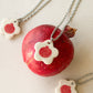 On top of a pomegranate lies a necklace with an off-white floral-shaped ceramic pendant featuring a red pomegranate drawing. Two similar necklaces are to the side.