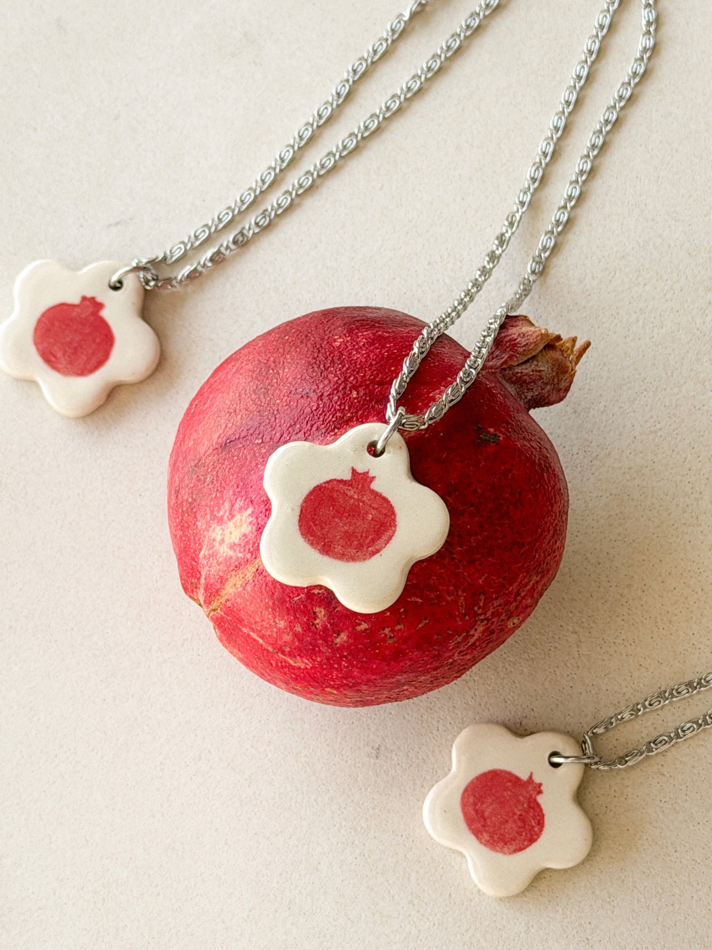 On top of a pomegranate lies a necklace with an off-white floral-shaped ceramic pendant featuring a red pomegranate drawing. Two similar necklaces are to the side.