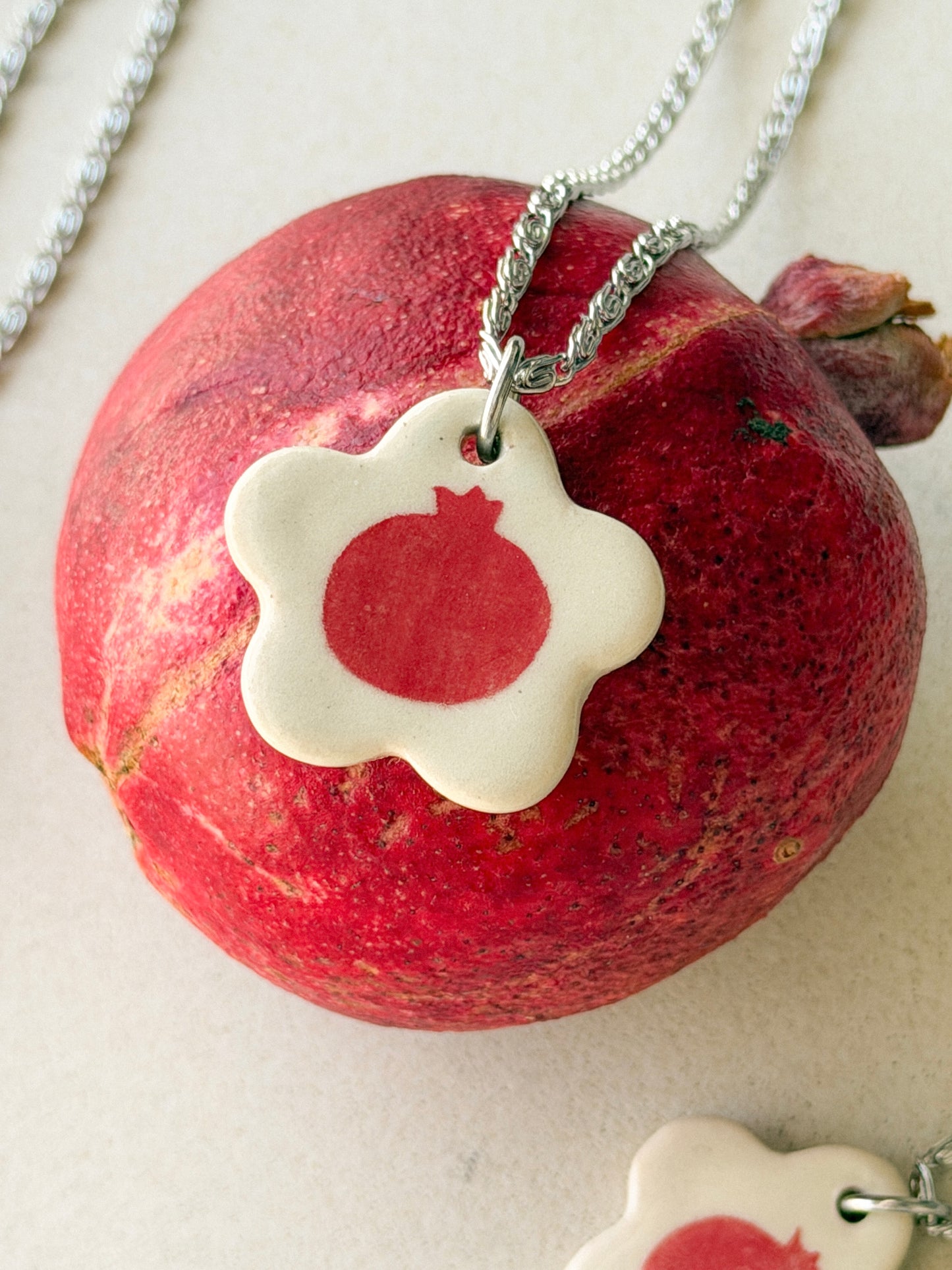 On top of a pomegranate lies a necklace with an off-white  floral-shaped ceramic pendant featuring a red pomegranate drawing