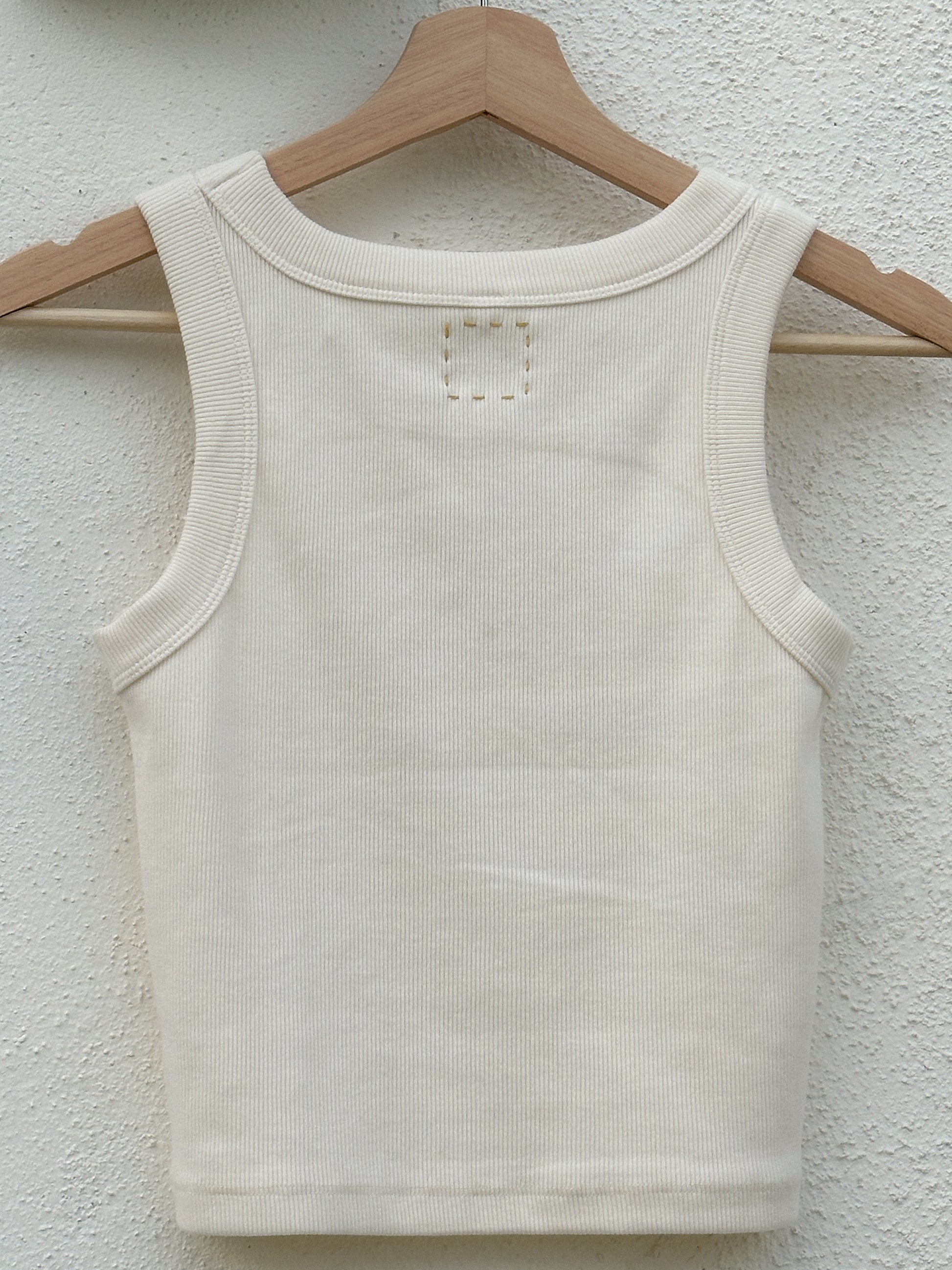 The back of a beige sleeveless tank top hanging on a wooden clothes hanger. The top of the tank shows a yellow hand-embroidered box