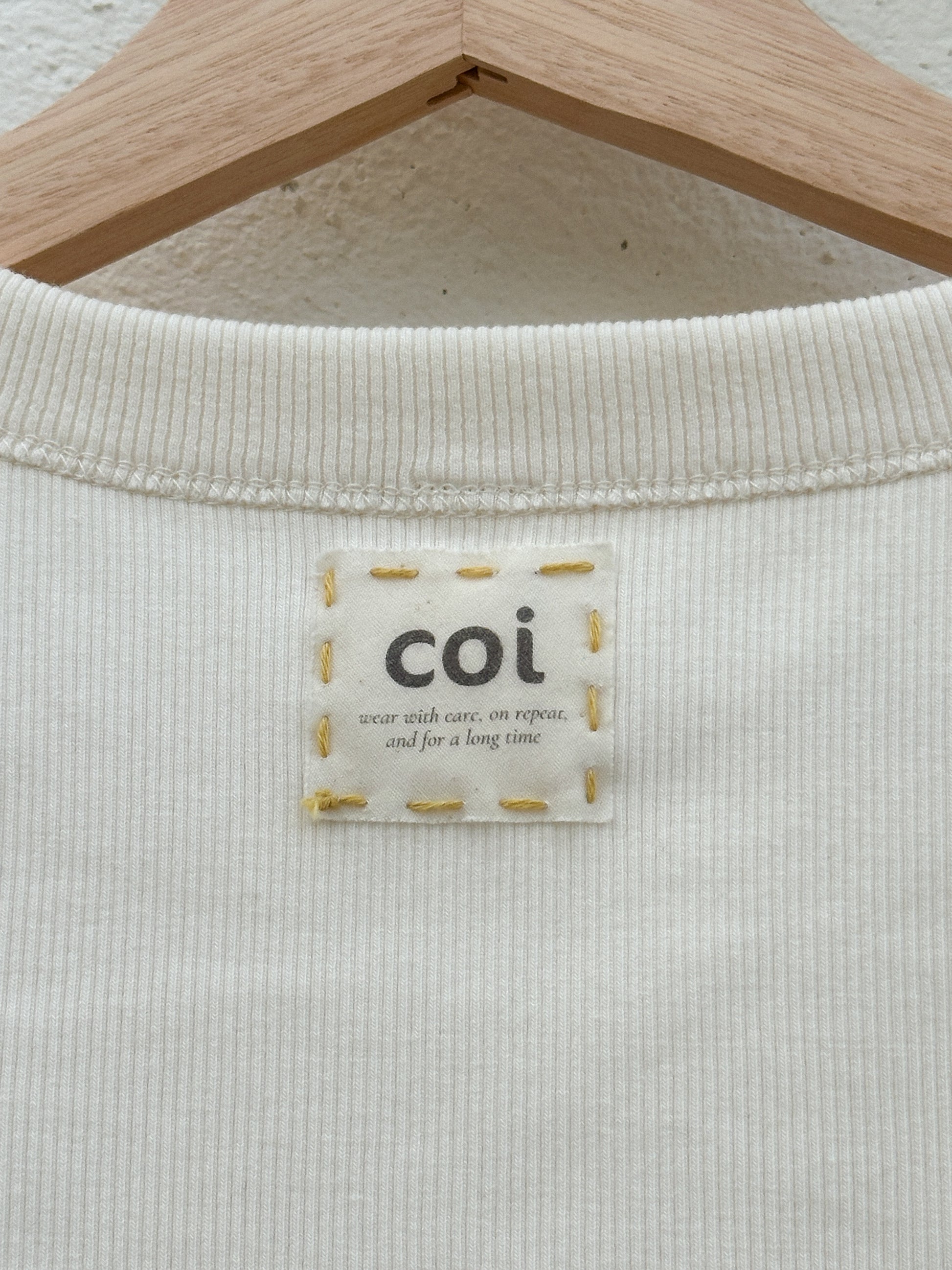 Up-close image of the inside of the beige tank top, showing a logo label for "coi" hand-embroidered on with yellow thread.