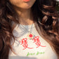 Close-up of person with long hair pouting, wearing a white baby tee with the Chinese words 寶寶 and "bao bao" on it and a necklace with a flower-shaped ceramic pendant featuring a pomegranate