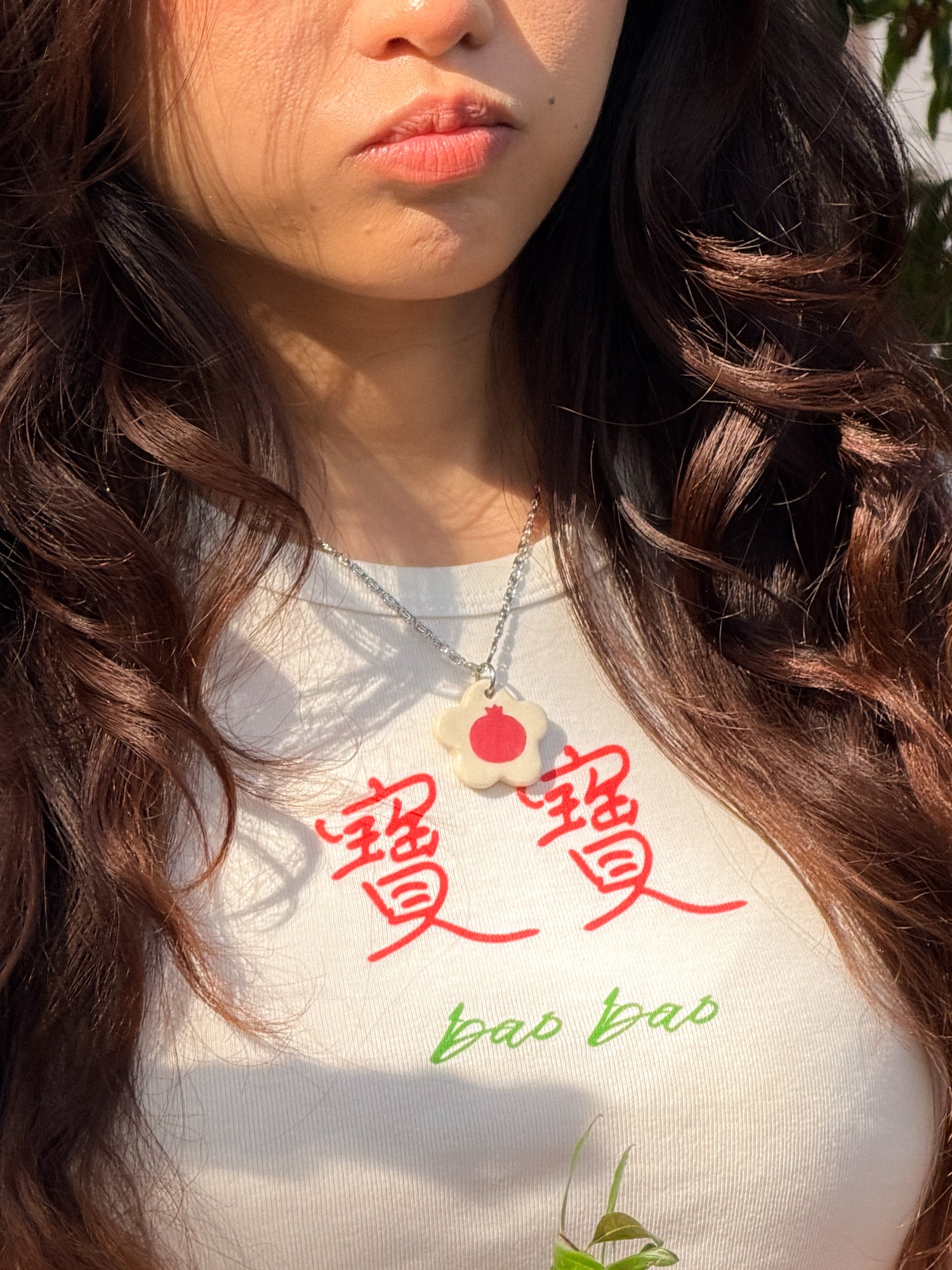 Close-up of person with long hair pouting, wearing a white baby tee with the Chinese words 寶寶 and "bao bao" on it and a necklace with a flower-shaped ceramic pendant featuring a pomegranate