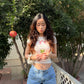 Woman with long hair wearing denim shorts and a white baby tee with the Chinese words 寶寶 and "bao bao" on it and holding pomegranate leaves in front of a pomegranate tree