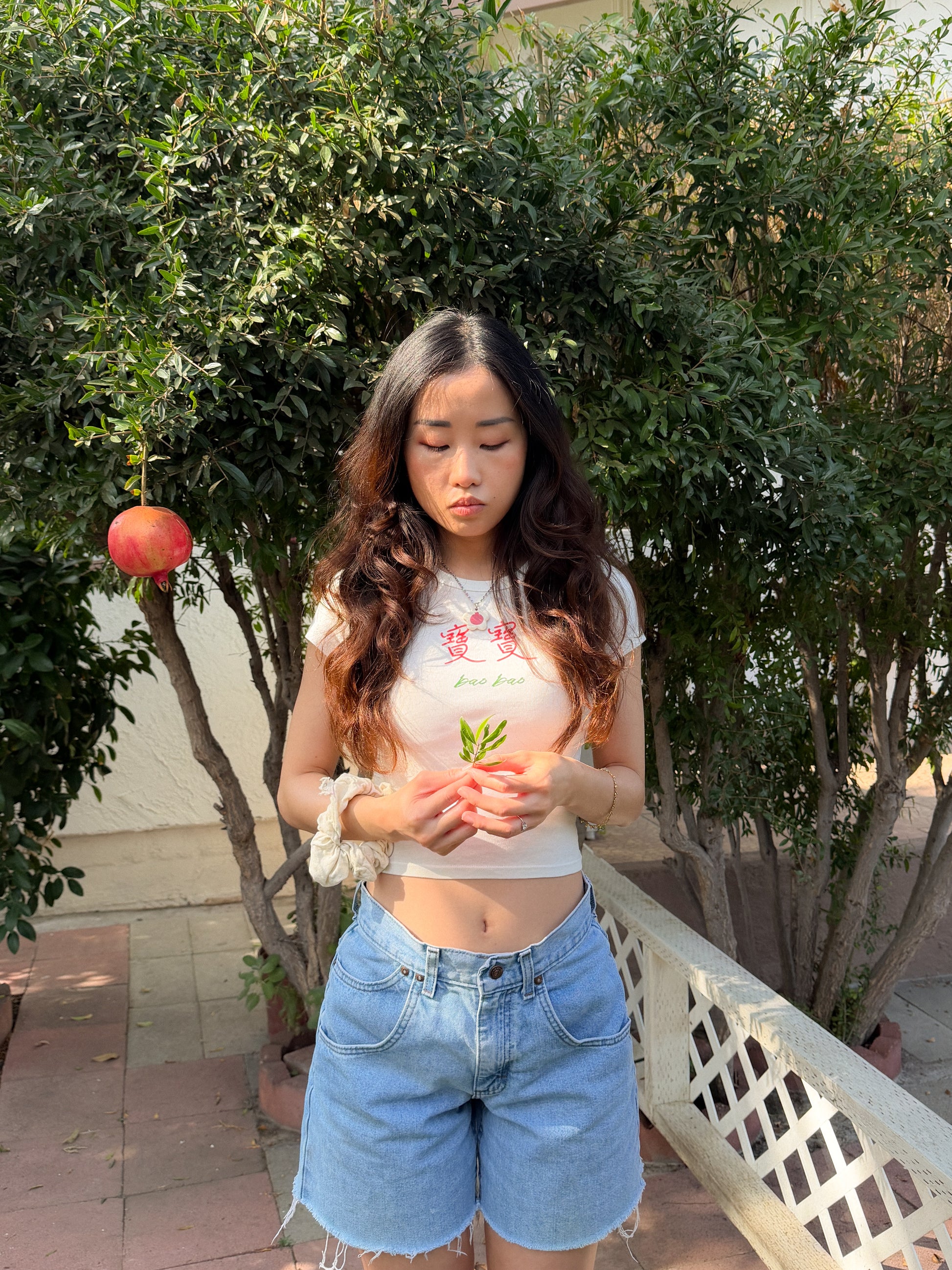 Woman with long hair wearing denim shorts and a white baby tee with the Chinese words 寶寶 and "bao bao" on it and holding pomegranate leaves in front of a pomegranate tree