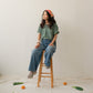 A Chinese Vietnamese woman with long hair wearing a green t-shirt and blue jeans sits on a wooden chair with her face turned slightly to the side and looking into the distance. On the floor are tangerines and Chinese broccoli on top of a white sheet.