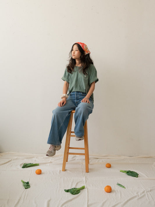 A Chinese Vietnamese woman with long hair wearing a green t-shirt and blue jeans sits on a wooden chair with her face turned slightly to the side and looking into the distance. On the floor are tangerines and Chinese broccoli on top of a white sheet.