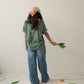A Chinese Vietnamese woman with long hair wearing a green t-shirt and blue jeans and holding Chinese broccoli in one hand stands with her back to the camera. On the floor are tangerines and Chinese broccoli on top of a white sheet.