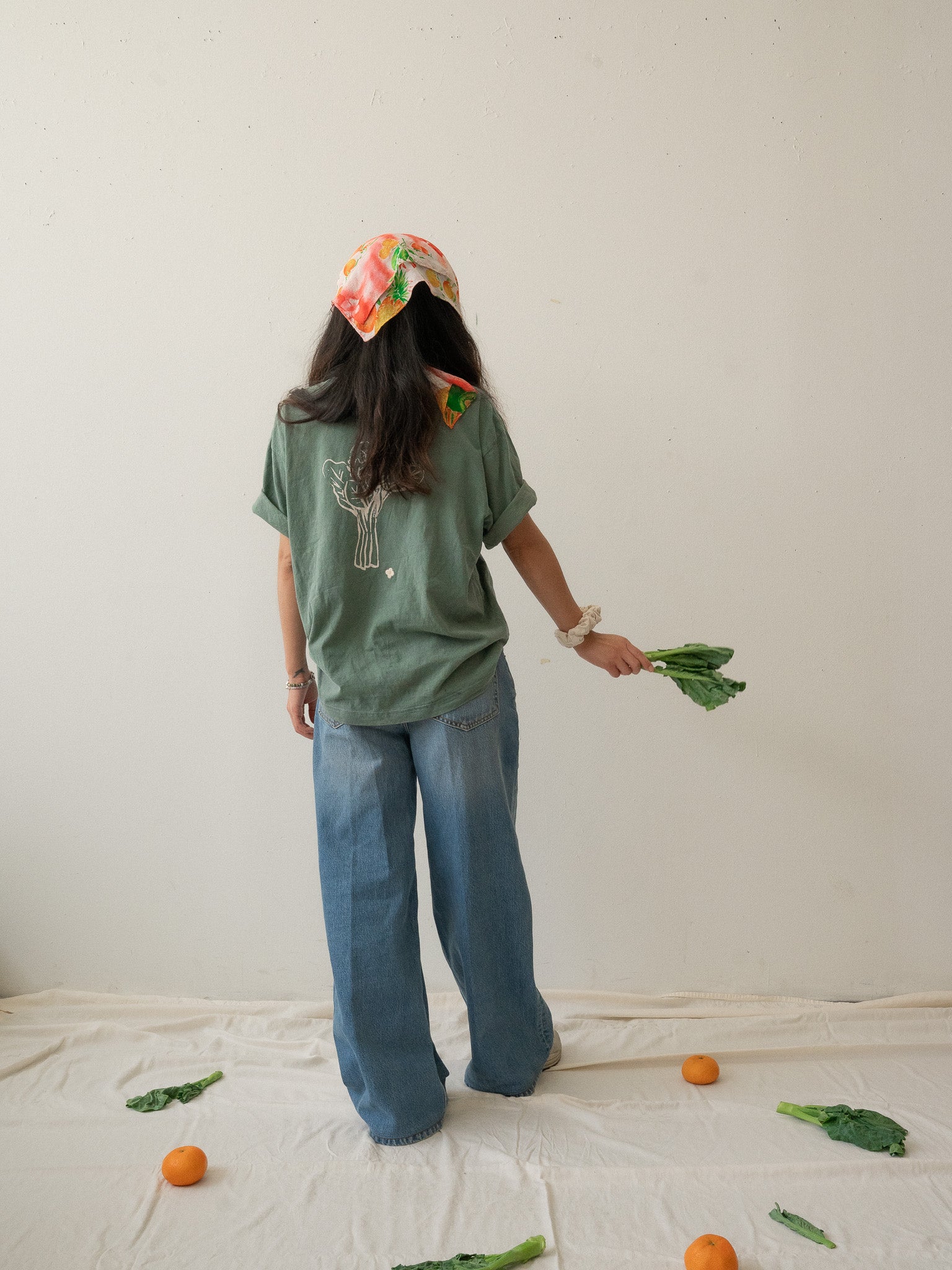 A Chinese Vietnamese woman with long hair wearing a green t-shirt and blue jeans and holding Chinese broccoli in one hand stands with her back to the camera. On the floor are tangerines and Chinese broccoli on top of a white sheet.