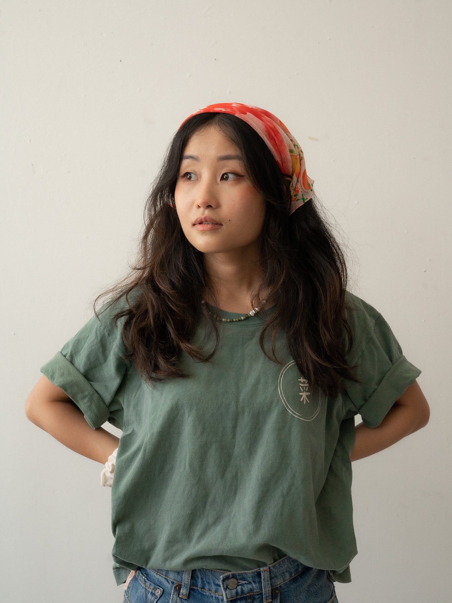 A Chinese Vietnamese woman with long hair wearing a green t-shirt and blue jeans with her face turned slightly to the side and looking into the distance. Her hands are on her waist.