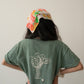 A Chinese Vietnamese woman wearing a green t-shirt with an illustration of a big Chinese broccoli and small embroidered white flower and blue jeans stands with her back to the camera. Her hands are on her waist.