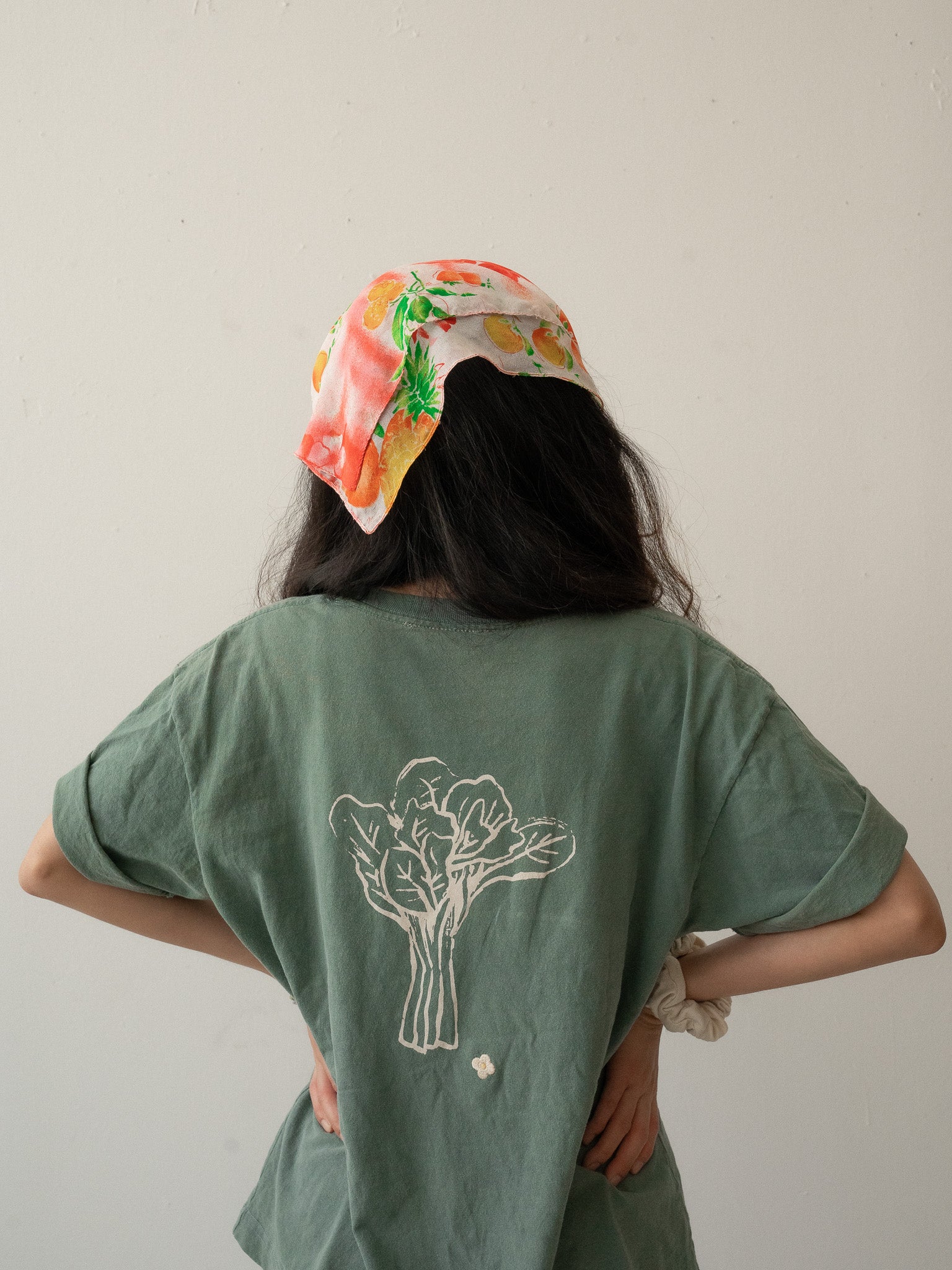 A Chinese Vietnamese woman wearing a green t-shirt with an illustration of a big Chinese broccoli and small embroidered white flower and blue jeans stands with her back to the camera. Her hands are on her waist.