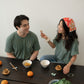 A Chinese man and Chinese Vietnamese woman wearing the same dark green shirt sit around a wooden table. Both are smiling, while one is holding a ha gow with her chopsticks. There are bowls of dim sum, tea cups, and a plate of Chinese broccoli on the table.