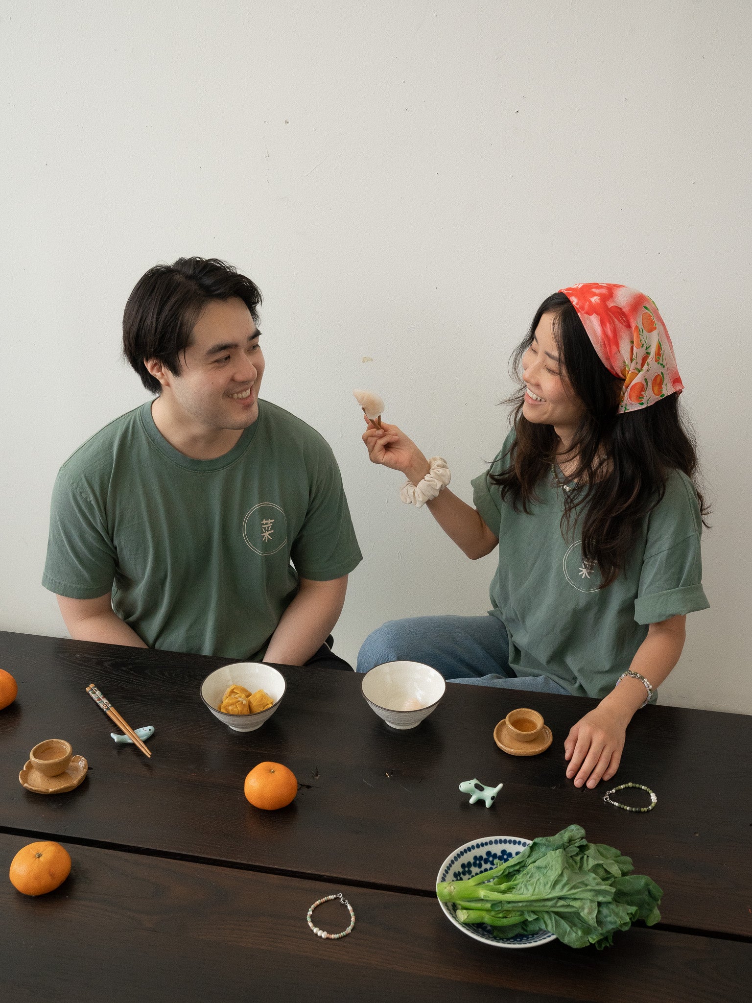 A Chinese man and Chinese Vietnamese woman wearing the same dark green shirt sit around a wooden table. Both are smiling, while one is holding a ha gow with her chopsticks. There are bowls of dim sum, tea cups, and a plate of Chinese broccoli on the table.