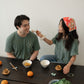 A Chinese man and Chinese Vietnamese woman wearing the same dark green shirt sit around a wooden table. Both are smiling, while one is holding up a ha gow close to the other’s face with her chopsticks. There are bowls of dim sum, tea cups, and a plate of Chinese broccoli on the table.