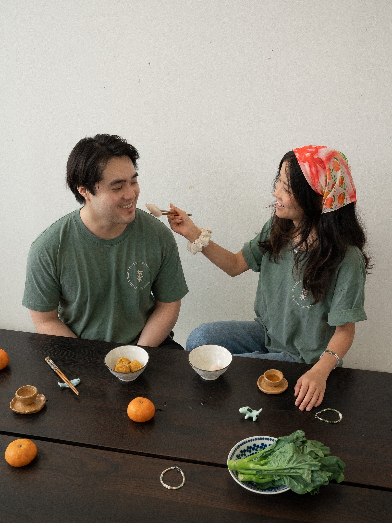 A Chinese man and Chinese Vietnamese woman wearing the same dark green shirt sit around a wooden table. Both are smiling, while one is holding up a ha gow close to the other’s face with her chopsticks. There are bowls of dim sum, tea cups, and a plate of Chinese broccoli on the table.