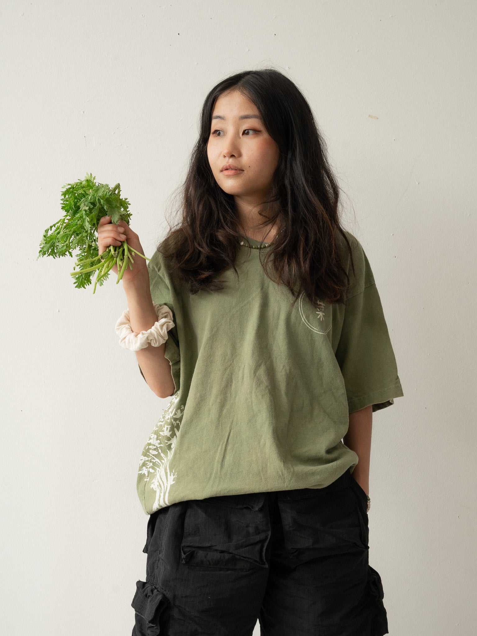 A Chinese Vietnamese woman wearing a yellow green t-shirt and black shorts stands with her back to the camera. She’s holding a bunch of tong ho in one hand.