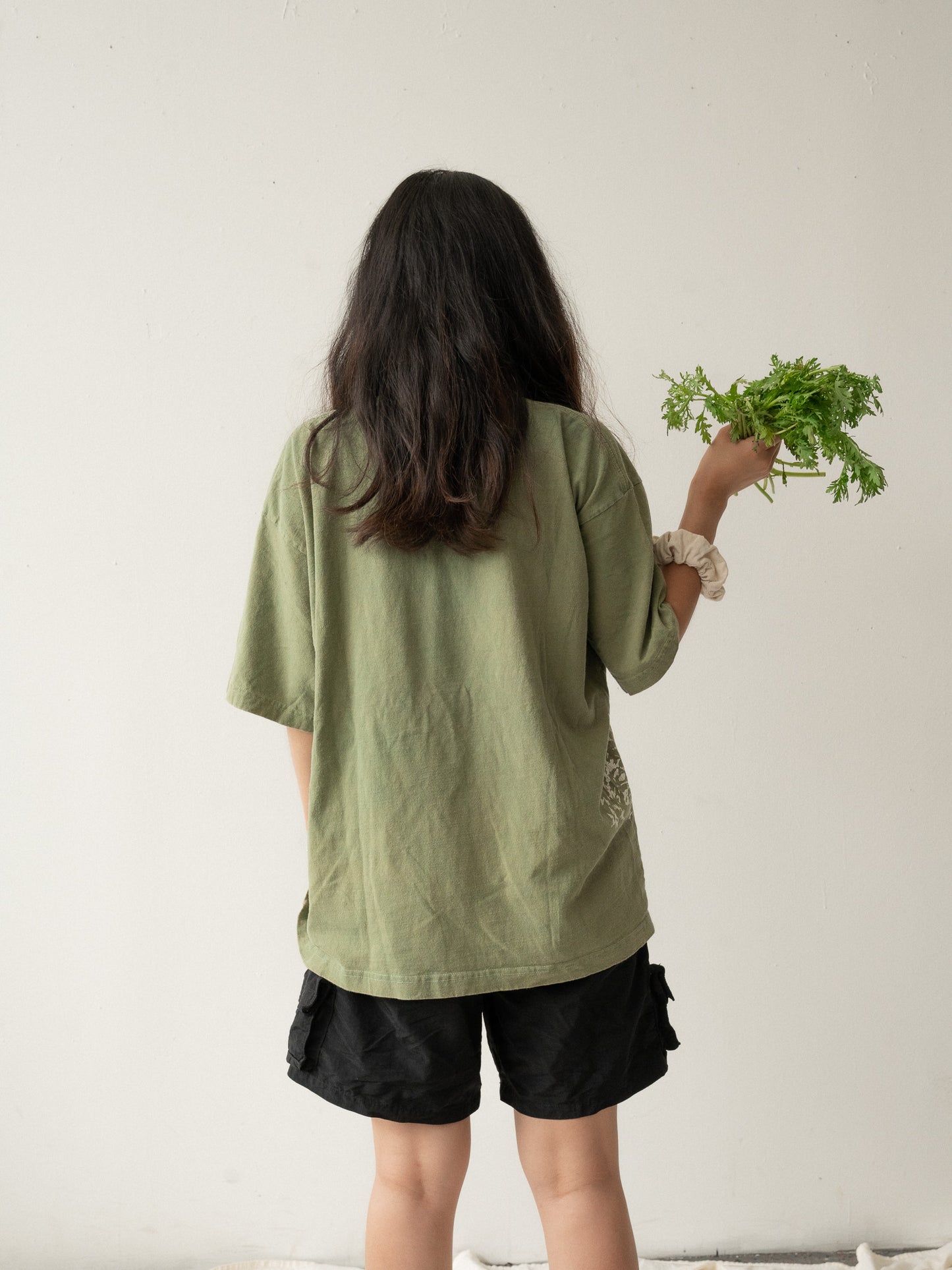 A Chinese Vietnamese woman wearing a yellow green t-shirt and black shorts stands with her back to the camera. She’s holding a bunch of tong ho in one hand.