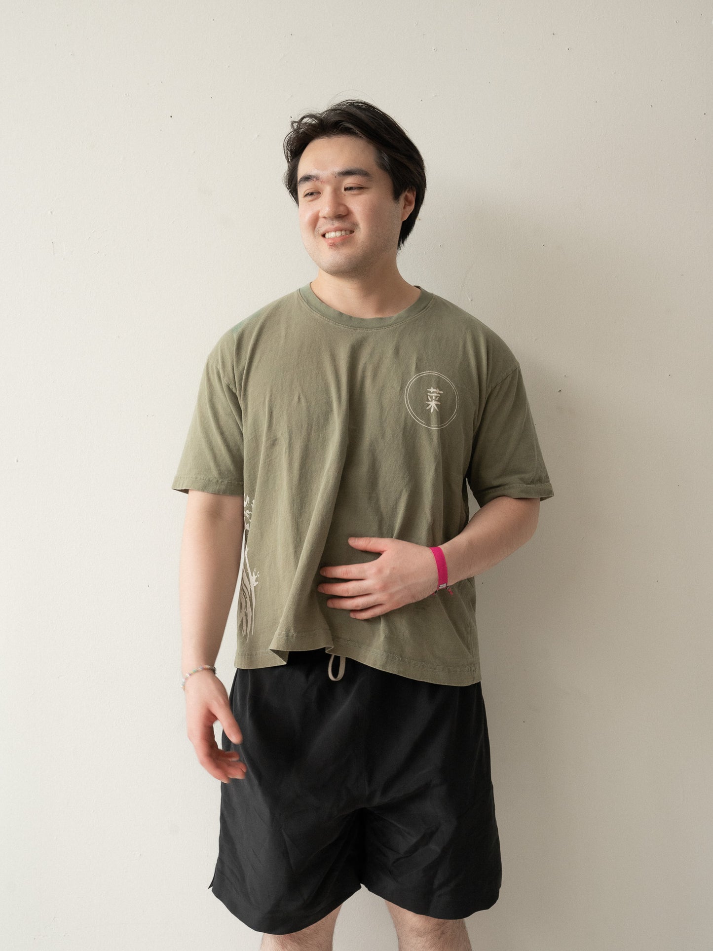 A Chinese man with short hair wearing a yellow brownish green t-shirt and black shorts with his face turned slightly to the side smiling and looking into the distance.