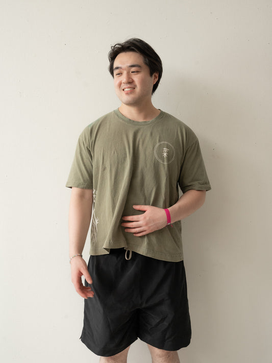 A Chinese man with short hair wearing a yellow brownish green t-shirt and black shorts with his face turned slightly to the side smiling and looking into the distance.